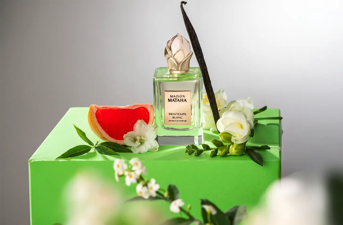 Een flacon Maison Mataha Printemps Blanc Extrait de Parfum staat op een felgroene doos, omringd door een halve grapefruit, een vanillestokje, witte bloemen en groene bladeren, met een lichte, frisse achtergrond.