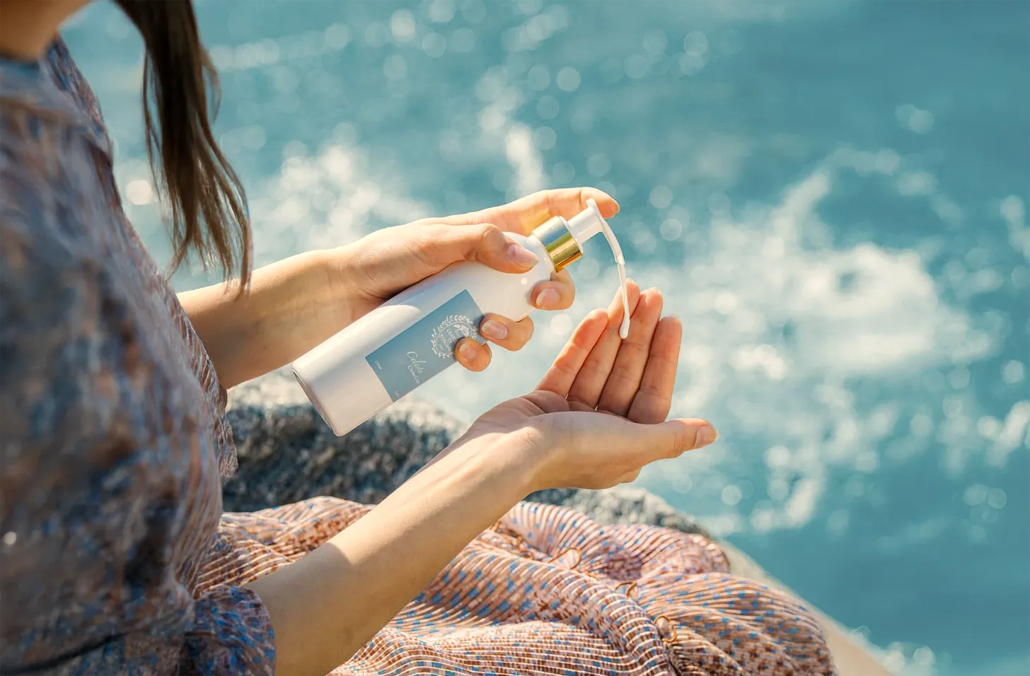 Vrouw pompt bodylotion van Giardini di Toscana Celeste uit een witte fles met gouden pomp op haar hand, zittend bij helderblauw water.